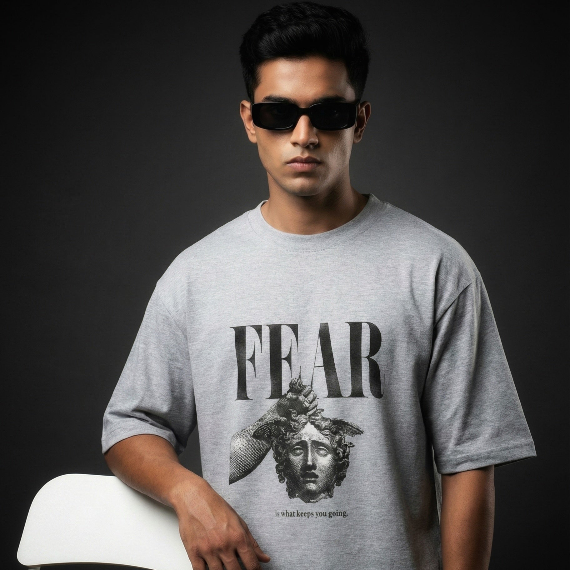 Man wearing a gray t-shirt with a graphic design and text, sitting on a white chair against a dark background.